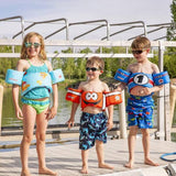 Three children wearing Airhead Elite Child Life Vests in different designs smile while standing on a dock by the water.